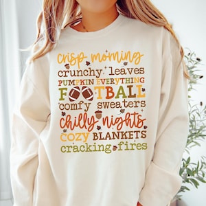 May include: Cream-colored sweatshirt with fall-themed text in various colors. The text includes phrases like "crisp morning," "crunchy leaves," "football," and "chilly nights." The design incorporates leaf and acorn graphics.