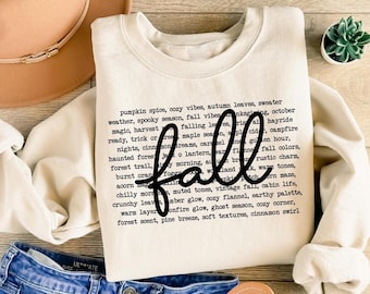 May include: Cream-colored sweatshirt with the word "fall" in a black script font. The sweatshirt is surrounded by fall-themed words like "pumpkin spice" and "autumn leaves". The sweatshirt is paired with blue jeans, sandals, and a brown hat.