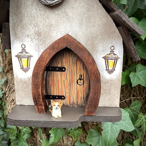 May include: A small birdhouse with a brown wooden roof and a white painted front. The front of the birdhouse has a small door with a brown wooden frame and a brown wooden door. There are two small lanterns painted on the front of the birdhouse. A small figurine of a dog is sitting on the porch of the birdhouse.