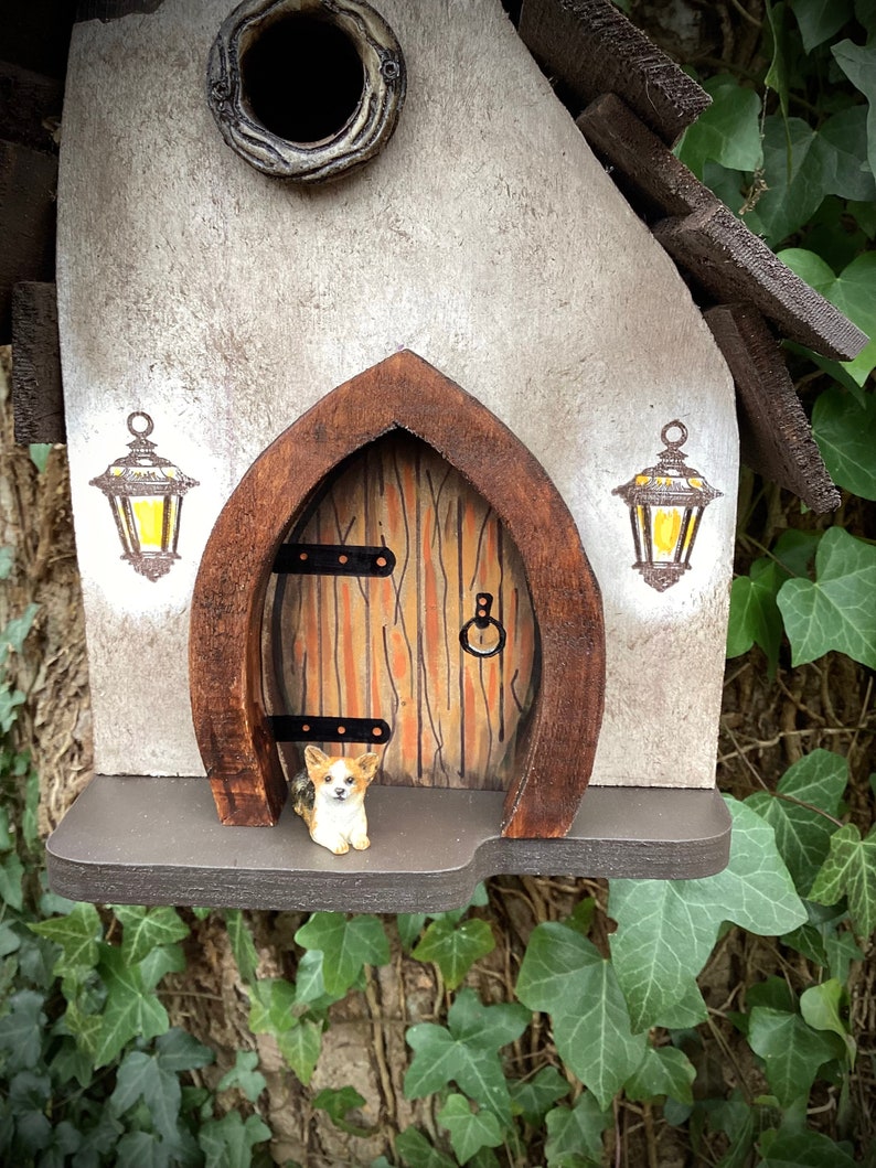 May include: A small birdhouse with a brown wooden roof and a white painted front. The front of the birdhouse has a small door with a brown wooden frame and a brown wooden door. There are two small lanterns painted on the front of the birdhouse. A small figurine of a dog is sitting on the porch of the birdhouse.