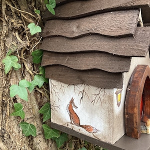 May include: A wooden birdhouse with a brown shingled roof and a painted scene of a fox on the side. The birdhouse is attached to a tree trunk with ivy growing on it.
