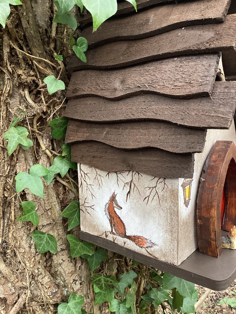 May include: A wooden birdhouse with a brown shingled roof and a painted scene of a fox on the side. The birdhouse is attached to a tree trunk with ivy growing on it.