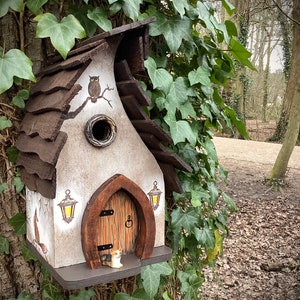 Könnte beinhalten: Ein fantasievolles Vogelhaus in Form eines Häuschens mit braunem Dach, weiß gestrichenem Äußeren und einer kleinen Tür. Das Vogelhaus hat zwei kleine Laternen an den Seiten und einen kleinen Eulen, der auf einem Ast über der Tür sitzt. Das Vogelhaus ist an einem Baumstamm befestigt, der mit grünem Efeu bedeckt ist.