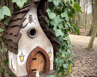 Puede incluir: Una casita para pájaros con forma de cabaña, con un techo marrón, un exterior pintado de blanco y una pequeña puerta. La casita tiene dos pequeñas linternas a los lados y un pequeño búho posado en una rama sobre la puerta. La casita está montada en un tronco de árbol cubierto de hiedra verde.