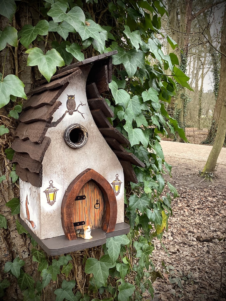 Wooden bird house featuring a crafty fox on the left, owl in the eaves, puppy dog waiting in the doorway, two glowing lanterns either side of the door, rabbits jumping and playing on the right. Curved wooden slatted roof. 30mm entrance hole. Stunning