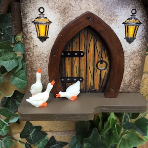 May include: A miniature wooden fairy house with a brown door and two yellow lanterns. Three white geese figurines are on the shelf below the door.