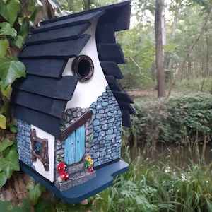 May include: A whimsical birdhouse shaped like a small cottage with a blue door, stone walls, and a black shingled roof. The birdhouse has a round entrance hole and a small window with a wooden frame. There are red and white mushrooms painted on the front of the birdhouse.