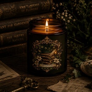 May include: A lit candle in a dark amber glass jar with a label featuring an ornate gold frame, a stack of books, a quill, and white flowers. The label text reads Literary Garden Society. The candle flame glows brightly within the glass container.