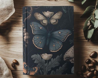 May include: A hardcover journal featuring a dark blue cover with a botanical design. The design includes illustrations of butterflies, flowers, and foliage in shades of blue, cream, and orange. The journal is surrounded by coffee beans and a linen cloth.
