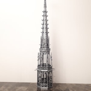 May include: A silver metal model of a tall, ornate church spire with a cross at the top. The spire is detailed with intricate carvings and arches.