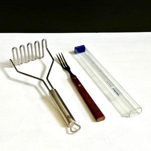 May include: A stainless steel potato masher and a fork with a wooden handle are displayed. A clear plastic case with a blue cap is also present. The potato masher has a looped head and a long handle. The fork has a dark brown handle.