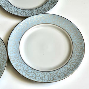 May include: Set of four white ceramic dinner plates with a light blue and gold decorative border. The border features a swirling, floral pattern.