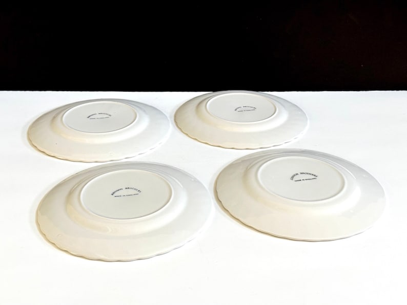 May include: Four white ceramic plates with a scalloped edge. The plates have a raised center and a small inscription on the bottom that reads "Avon Broadsides Made in England".