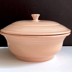 May include: A pale pink ceramic casserole dish with a lid. The dish has a rounded shape and a smooth, glossy finish. The lid has a small knob on top.