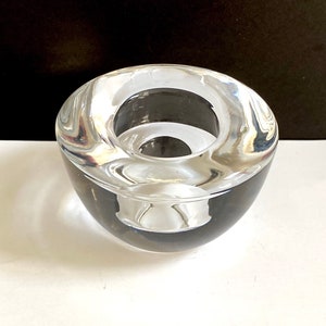 May include: A clear glass candle holder with a round, slightly curved design. The holder has a wide opening at the top and a smaller opening at the bottom.