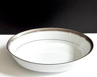 May include: White ceramic serving bowl with a silver rim and a thin black line. The bowl has a simple, elegant design, suitable for serving various dishes. The bowl is round and has a shallow depth.