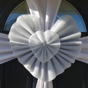 May include: A white tulle heart-shaped decoration with a silver tulle bow. The decoration is attached to a white fabric backdrop.