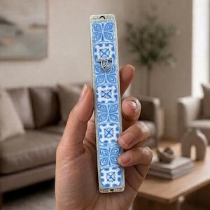 以下が含まれることがあります： 青と白の繰り返し花柄タイル模様が施された長方形のメズーザケース。細長い形状で、銀色の金属フレームが特徴です。前面中央には銀色のヘブライ文字「シン」が配置されており、装飾的なデザインとなっています。