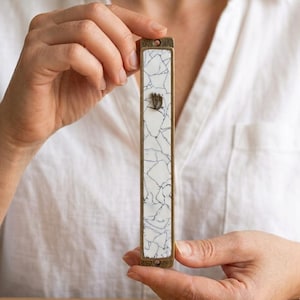 May include: A rectangular mezuzah case featuring a white surface with a black crackle pattern. The case has a metallic bronze-toned frame with mounting holes at the top and bottom. A small, raised silver-colored Hebrew letter Shin is centered.