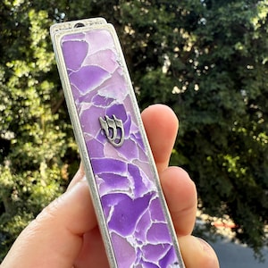 May include: A rectangular mezuzah case with a silver-colored frame and a mosaic-like design in shades of purple. A silver-colored Hebrew letter is visible on the front. The case is held in a person's hand.