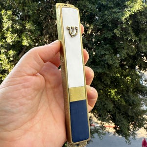 May include: A rectangular mezuzah case with a gold-colored metal frame. The case features a white center section, a gold band, and a navy blue bottom. A Hebrew letter is visible on the white section. The background is blurred.