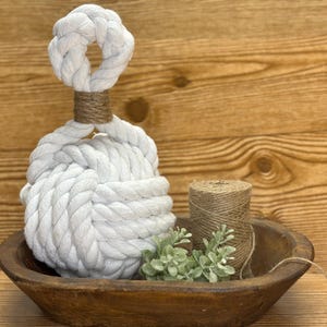 May include: A decorative white rope knot, with a smaller knot at the top, displayed in a wooden bowl. A spool of twine and greenery are also in the bowl. The background is a wooden surface.