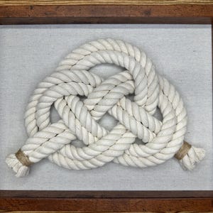 Op de afbeelding: Een ingelijst nautisch kunstwerk met een witte touwknoop tegen een lichtgrijze achtergrond. Het touw is strak geweven tot een decoratieve knoop en de lijst is gemaakt van donkerbruin hout. Het kunstwerk is een decoratief stuk.
