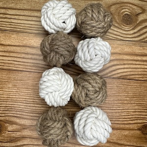 May include: Eight decorative nautical rope balls in alternating white and brown colors. The balls are arranged in two rows of four, showcasing their intricate knotwork. The background is a wooden surface with visible grain.
