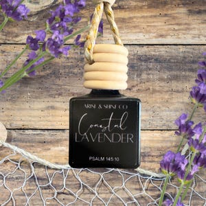 May include: A black air freshener with a wooden top and a rope hanger. The label reads "Coastal Lavender" with the text "Psalm 145:10" below. Lavender sprigs and a fishing net are in the background.