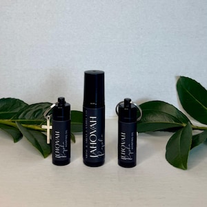 May include: Three black bottles of anointing oil. The bottles are labeled "JEHOVAH Rapha" and "Aointing Oil". One bottle has a small silver cross attached to it. The bottles are arranged in front of green leaves.