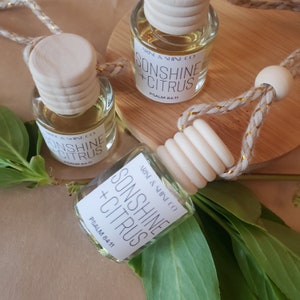 May include: Three small glass bottles of air freshener with wooden caps and hanging cords. Each bottle has a label that reads "SONSHINE + CITRUS" and "PSALM 84:11". The bottles are surrounded by green leaves and sit on a light brown surface.