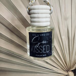 May include: Clear glass car air freshener with a white wooden top and a braided rope hanger. The label reads "Son Kissed" and "Psalm 34:5". The air freshener is hanging in front of a dried palm leaf.