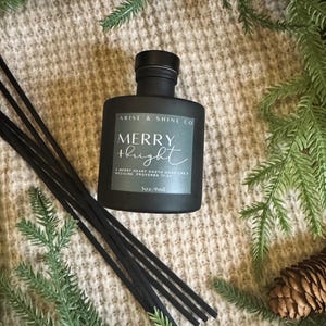 May include: A black diffuser bottle with the text "MERRY + bright" and the brand name "ARISE & SHINE CO". The bottle contains 3oz/9ml of liquid and is surrounded by black reeds and evergreen branches.