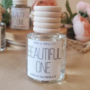 May include: A clear glass bottle with a white label that reads "ARISE & SHINE CO. BEAUTIFUL ONE SONG OF SOLOMON 2:13". The bottle has a wooden cap with a gold string.