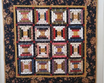 May include: A black and gold quilt with a patchwork design featuring 16 squares of different fabrics. Each square is made up of multiple strips of fabric in various colors and patterns. The quilt is framed with a black and gold border.