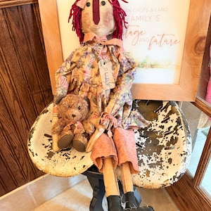 May include: A handmade doll with red yarn hair, wearing a floral dress and pink shorts, sits on a distressed metal stool. The doll has black boots and a tag that reads "RUBY". A small teddy bear is nestled beside the doll. A framed picture is in the background.