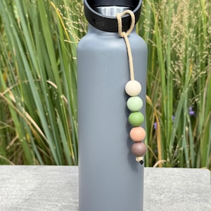 May include: A grey insulated water bottle with a black cap and handle. A beige cord is attached to the handle, decorated with five colourful beads in white, green, peach, and brown. The bottle stands upright against green foliage.