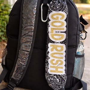 Op de afbeelding: Zwarte rugzak met een gewatteerde schouderband met zilveren glitter en een decoratieve band met de woorden "GOLD RUSH" in goudglitter. De decoratieve band heeft een zwart-wit paisley patroon.