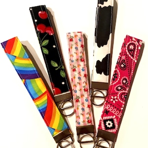 May include: Five colorful keychains with different patterns. The keychains are made of fabric and have metal rings. The patterns include a rainbow, cherries, pink flowers, cow print, and a pink bandana.