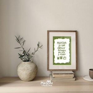 May include: A framed print with a green background and white text that reads "Matcha is the only green flag I need". There are three green tea cup illustrations below the text. The print is sitting on a wooden surface with a vase of green plants and a stack of books.