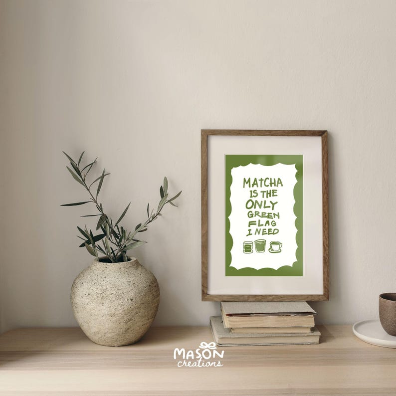 May include: A framed print with a green background and white text that reads "Matcha is the only green flag I need". There are three green tea cup illustrations below the text. The print is sitting on a wooden surface with a vase of green plants and a stack of books.