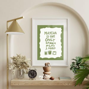 May include: A green and white framed print with the text "Matcha is the only green flag I need" and three green tea cup illustrations. The print is hanging on a white wall above a wooden shelf with a small elephant figurine, a clock, and a vase with dried flowers.