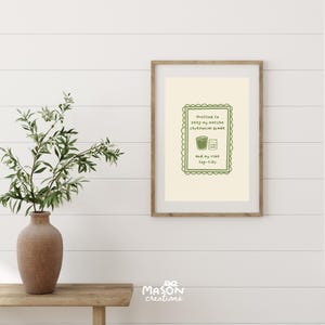 May include: A framed print with a pale yellow background and green text that reads "Hustling to keep my matcha ceremonial grade and my vibe top-tier". The text is surrounded by a green decorative border. The print is in a wooden frame.