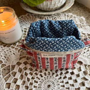 May include: A small, open fabric container with a red and white striped exterior and a blue floral interior. The container has a zipper closure and a tag that reads "Moon Altered Atelier".
