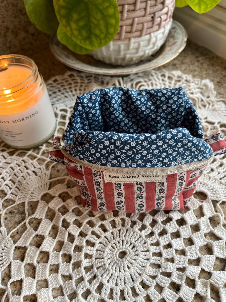 May include: A small, open fabric container with a red and white striped exterior and a blue floral interior. The container has a zipper closure and a tag that reads "Moon Altered Atelier".