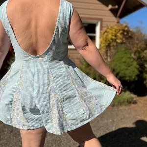 May include: Light blue and white striped romper with a low back and floral patterned panels. The romper has a flared skirt and a scoop neckline. The fabric appears to be seersucker.