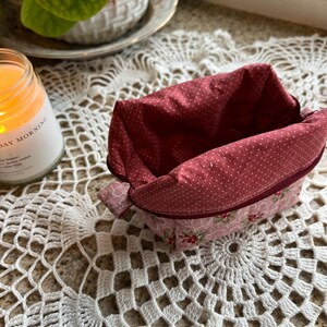 May include: A small, open fabric basket with a red polka dot interior and a pink floral exterior. The basket sits on a white crocheted doily. A lit candle and a potted plant are in the background.