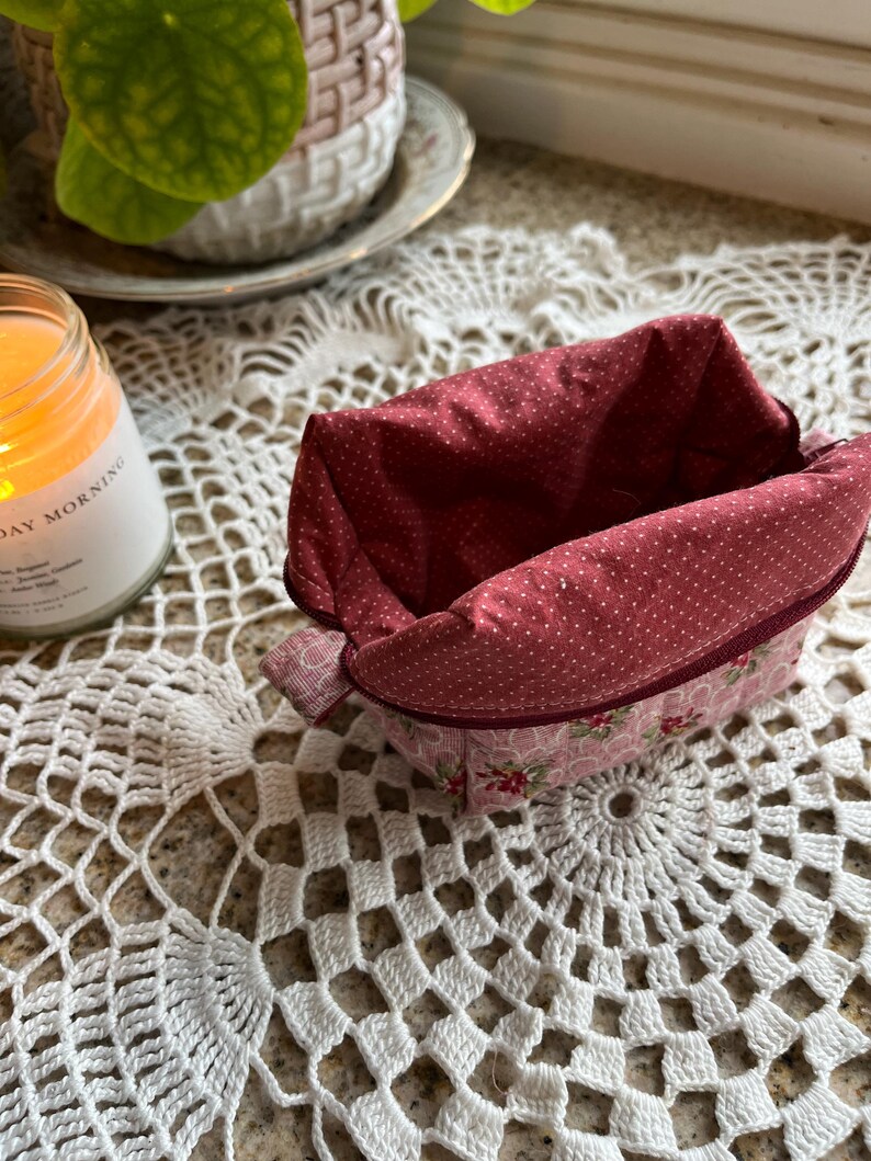 May include: A small, open fabric basket with a red polka dot interior and a pink floral exterior. The basket sits on a white crocheted doily. A lit candle and a potted plant are in the background.