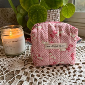 May include: A small, quilted pink pouch with a repeating pattern of white and pink dog illustrations. The pouch has a zipper closure and a small tag that reads "Moon Altered Atelier". A lit candle and a houseplant are in the background.