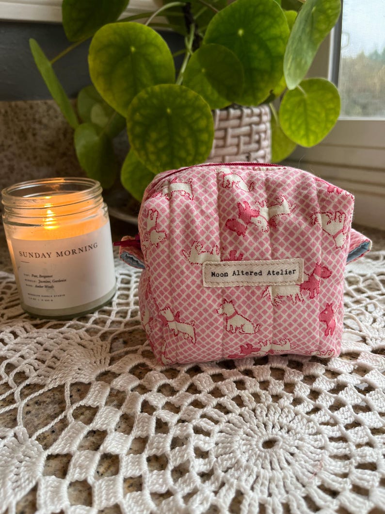 May include: A small, quilted pink pouch with a repeating pattern of white and pink dog illustrations. The pouch has a zipper closure and a small tag that reads "Moon Altered Atelier". A lit candle and a houseplant are in the background.